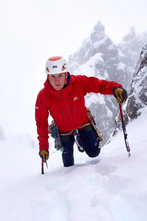 Ein Bergsteiger klettert mit Eispickeln durch eine verschneite, felsige Berglandschaft.