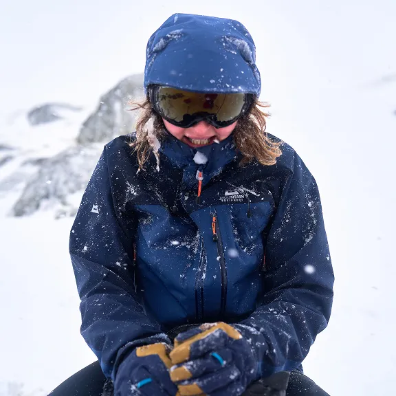 Eine Person sitzt im Schneetreiben und trägt eine dunkelblaue Outdoorjacke und eine Schneebrille.