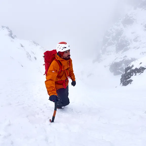 Ein Bergkletterer in oranger Jacke und weißem Helm geht mit Eispickel durch eine steile, verschneite Berglandschaft. 